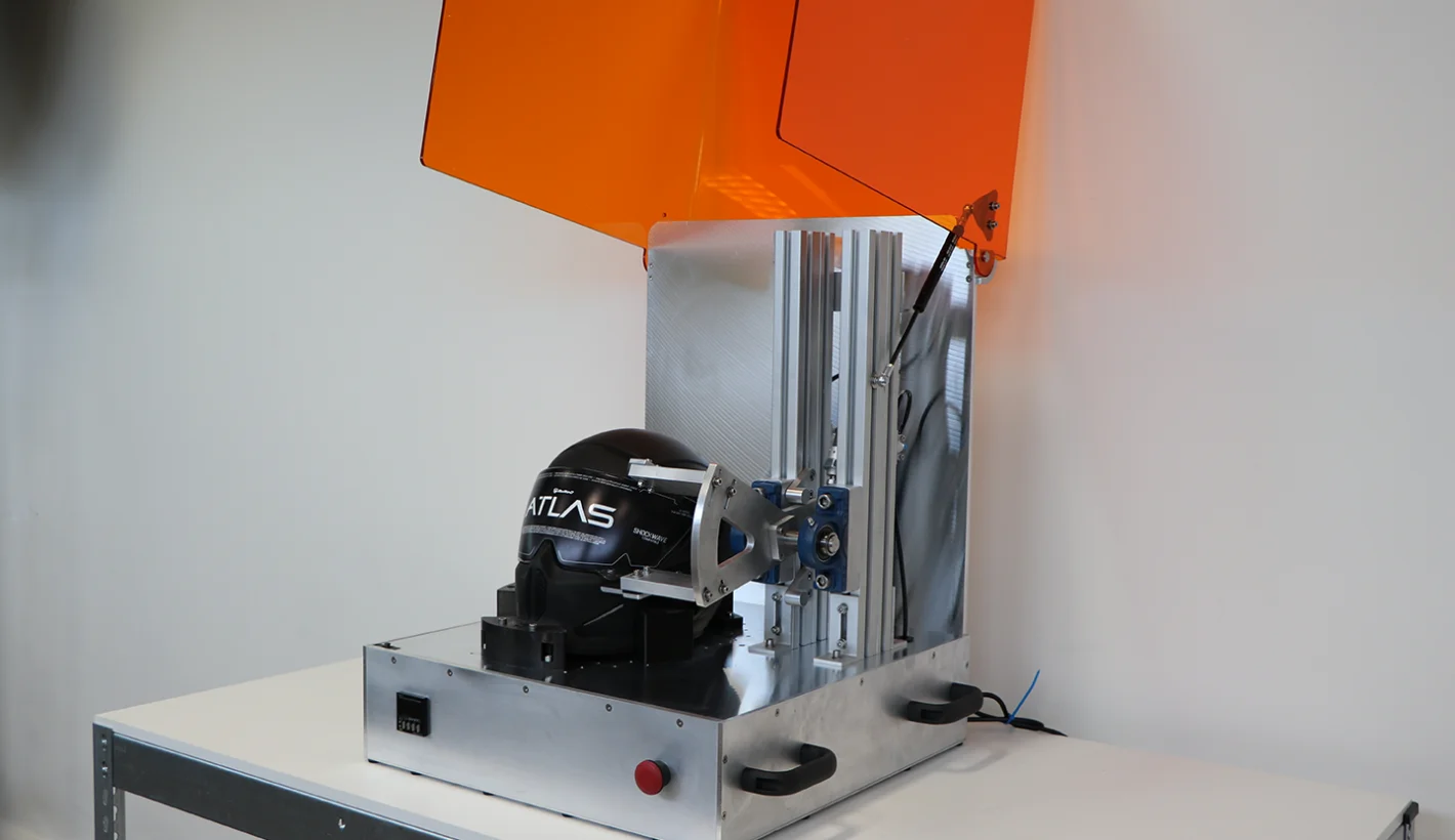 Black motorcycle helmet clamped into visor test rig with orange perspex box lid lifted