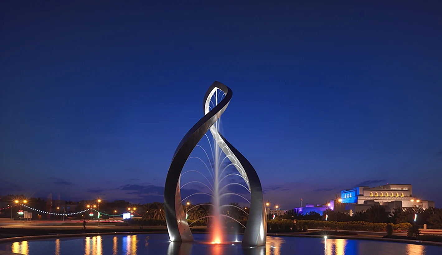 Side angle of Arches of Oman sculpture in water in Oman at night time surrounded by city lights